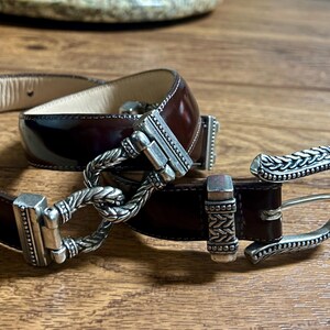 Puede incluir: Un cintur&oacute;n de cuero marr&oacute;n con una hebilla plateada. La hebilla tiene un dise&ntilde;o trenzado y un cierre decorativo.