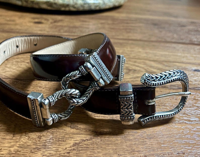 Puede incluir: Un cintur&oacute;n de cuero marr&oacute;n con una hebilla plateada. La hebilla tiene un dise&ntilde;o trenzado y un cierre decorativo.