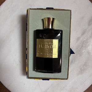 May include: A vintage black glass bottle of "Bath Oil" by Est&eacute;e Lauder, with a gold-colored cap and label, presented in a rectangular box with a gold and blue design. The bottle is displayed on a white marble surface.
