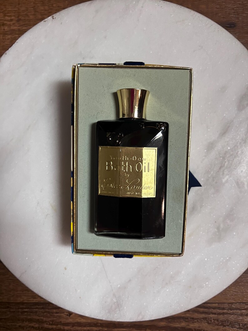 May include: A vintage black glass bottle of "Bath Oil" by Est&eacute;e Lauder, with a gold-colored cap and label, presented in a rectangular box with a gold and blue design. The bottle is displayed on a white marble surface.