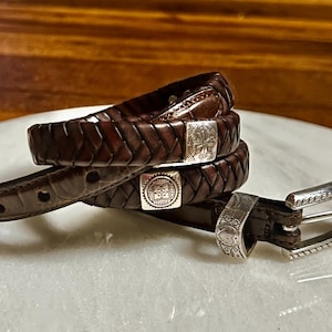 K&ouml;nnte beinhalten: Ein brauner Lederg&uuml;rtel mit geflochtenem Design und silberfarbener Schnalle. Der G&uuml;rtel ist aufgerollt und zeigt das komplizierte Gewebe und die dekorativen Metallakzente. Die Schnalle hat ein verziertes Design.