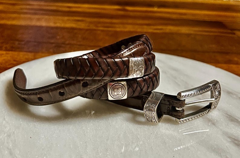 K&ouml;nnte beinhalten: Ein brauner Lederg&uuml;rtel mit geflochtenem Design und silberfarbener Schnalle. Der G&uuml;rtel ist aufgerollt und zeigt das komplizierte Gewebe und die dekorativen Metallakzente. Die Schnalle hat ein verziertes Design.