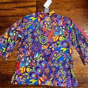 May include: A long-sleeved top with a vibrant, colorful floral and paisley pattern in shades of blue, purple, pink, yellow, and red. The top has a high neckline and a relaxed fit. The sleeves have a pink stripe down the side.