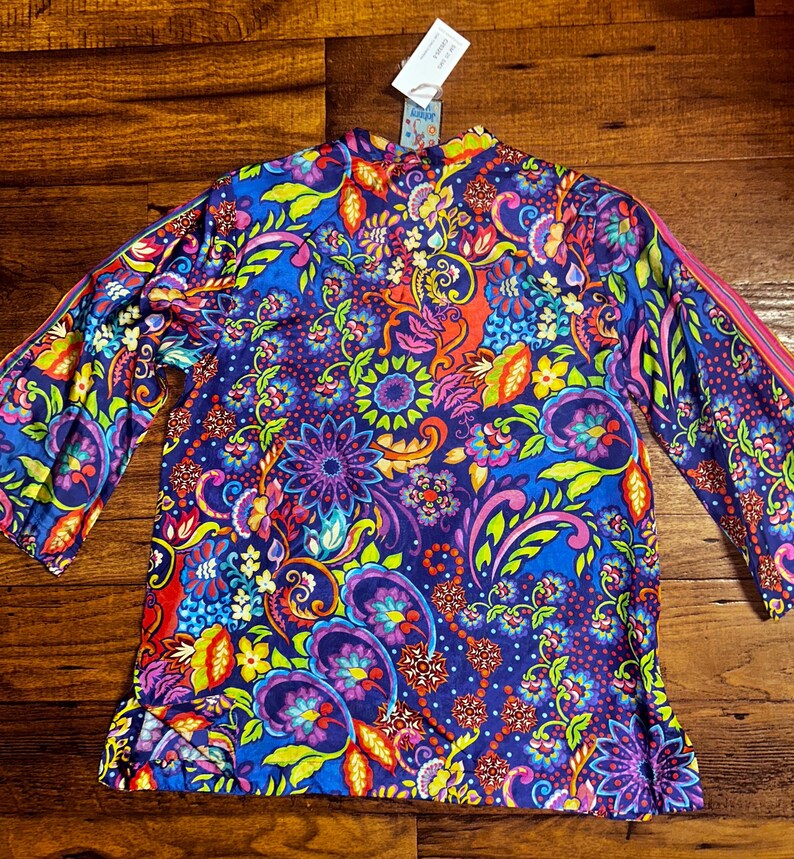 May include: A long-sleeved top with a vibrant, colorful floral and paisley pattern in shades of blue, purple, pink, yellow, and red. The top has a high neckline and a relaxed fit. The sleeves have a pink stripe down the side.