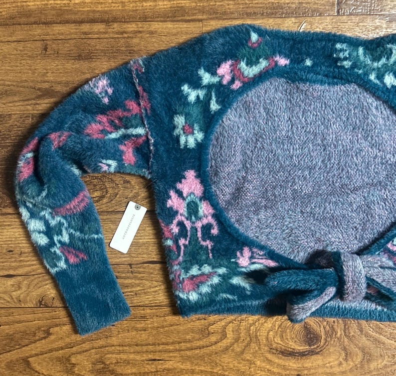May include: A teal blue fuzzy knit sweater with a pink and green floral pattern. The sweater has a large open back with a tie at the bottom.