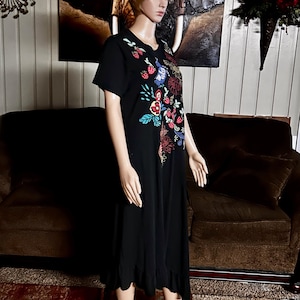 Puede incluir: Un vestido midi negro de manga corta con un vibrante patr&oacute;n bordado de flores y fresas en la parte superior delantera. La prenda tiene un cuello redondo y un dobladillo con volantes, exhibido en un maniqu&iacute; para mostrar su silueta.