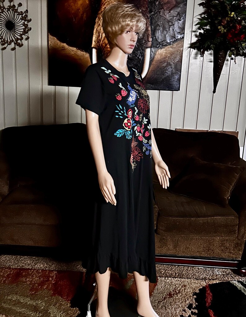 Puede incluir: Un vestido midi negro de manga corta con un vibrante patr&oacute;n bordado de flores y fresas en la parte superior delantera. La prenda tiene un cuello redondo y un dobladillo con volantes, exhibido en un maniqu&iacute; para mostrar su silueta.
