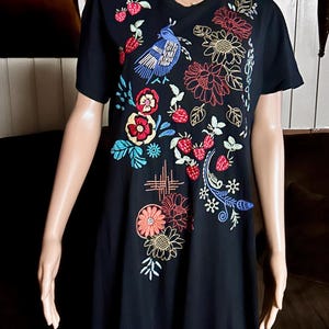 Puede incluir: Un vestido negro de manga corta con bordados coloridos de un p&aacute;jaro azul, fresas rojas y varios patrones florales en tonos rojos, amarillos y azules. El dise&ntilde;o se concentra en la parte frontal de la prenda, mostrando costuras detalladas.