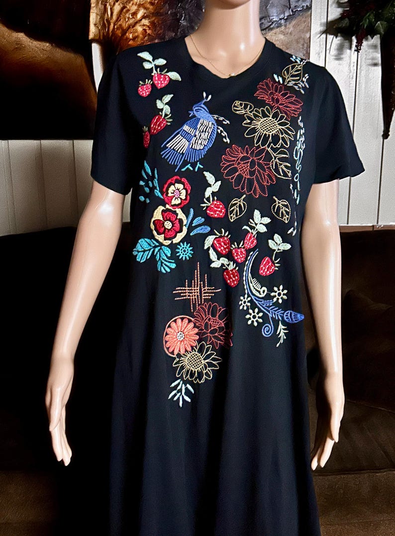 Puede incluir: Un vestido negro de manga corta con bordados coloridos de un p&aacute;jaro azul, fresas rojas y varios patrones florales en tonos rojos, amarillos y azules. El dise&ntilde;o se concentra en la parte frontal de la prenda, mostrando costuras detalladas.