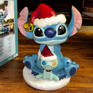 May include: A blue Stitch figurine wearing a red Santa hat with white trim and a gold bell. A smaller Stitch figure with a red scarf and Santa hat sits on a white base. The figurine is a holiday decoration.