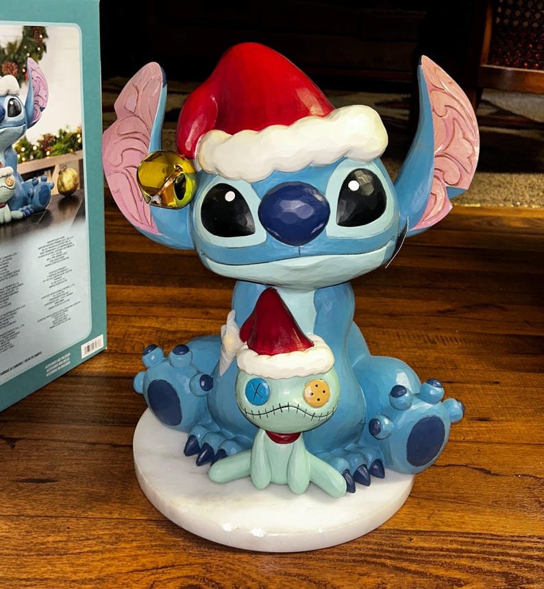 May include: A blue Stitch figurine wearing a red Santa hat with white trim and a gold bell. A smaller Stitch figure with a red scarf and Santa hat sits on a white base. The figurine is a holiday decoration.