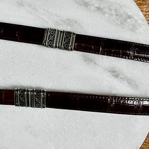 May include: Two brown leather belts with silver-colored metal accents. The belts have a crocodile skin texture and decorative metal clasps. The belts are displayed on a white marble surface.