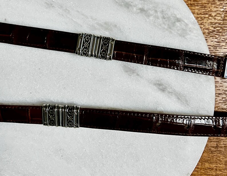 May include: Two brown leather belts with silver-colored metal accents. The belts have a crocodile skin texture and decorative metal clasps. The belts are displayed on a white marble surface.