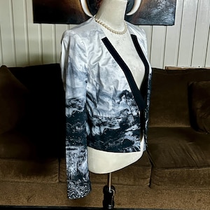 May include: A white and gray landscape print blazer with black lapels. The blazer is displayed on a mannequin. The landscape design features trees and a waterfall. The blazer has long sleeves.