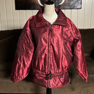 May include: A burgundy-colored jacket with a high collar and a zip-up front. The jacket has a belted waist and is displayed on a mannequin. The jacket is made of a shiny material, and the overall style is vintage.