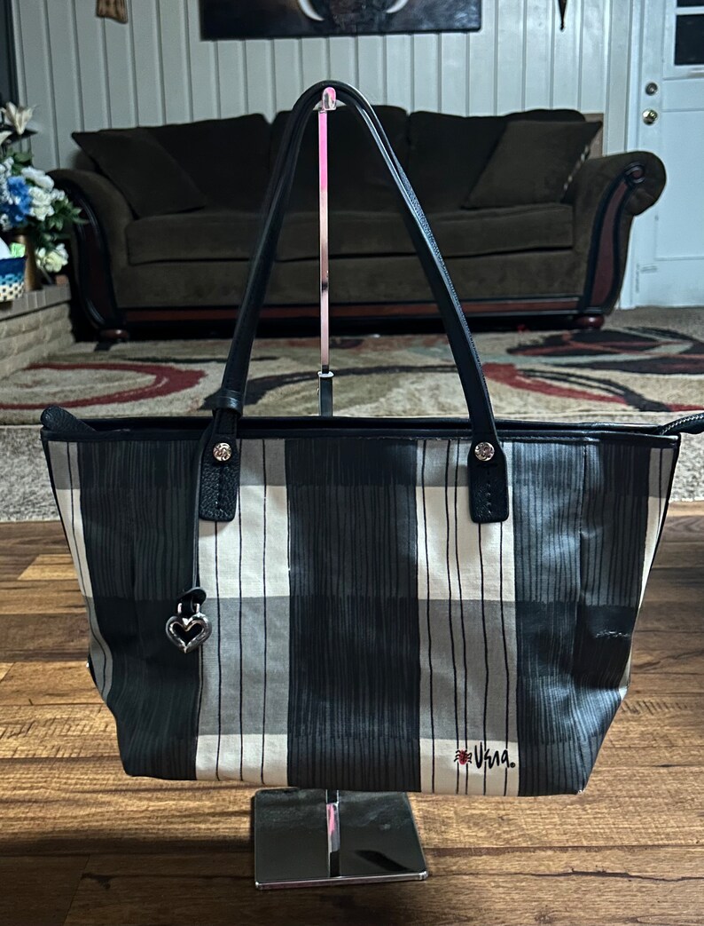 May include: A black and white plaid tote bag with black leather handles and a silver heart charm. The bag has the word "Vera" written on the bottom in red.