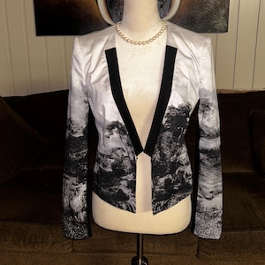 May include: A white blazer with a black and white landscape print. The blazer has black lapels and is displayed on a mannequin. A pearl necklace is around the mannequin's neck.