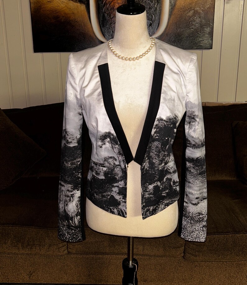 May include: A white blazer with a black and white landscape print. The blazer has black lapels and is displayed on a mannequin. A pearl necklace is around the mannequin's neck.