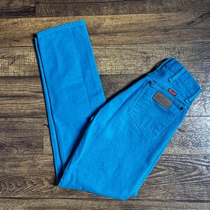 K&ouml;nnte beinhalten: Helle t&uuml;rkisfarbene Wrangler-Jeans. Die Jeans haben ein klassisches F&uuml;nf-Taschen-Design mit einem braunen Lederaufn&auml;her auf der Ges&auml;&szlig;tasche. Das Wrangler-Logo ist auf dem Aufn&auml;her sichtbar. Die Jeans liegen flach auf einer Holzoberfl&auml;che.