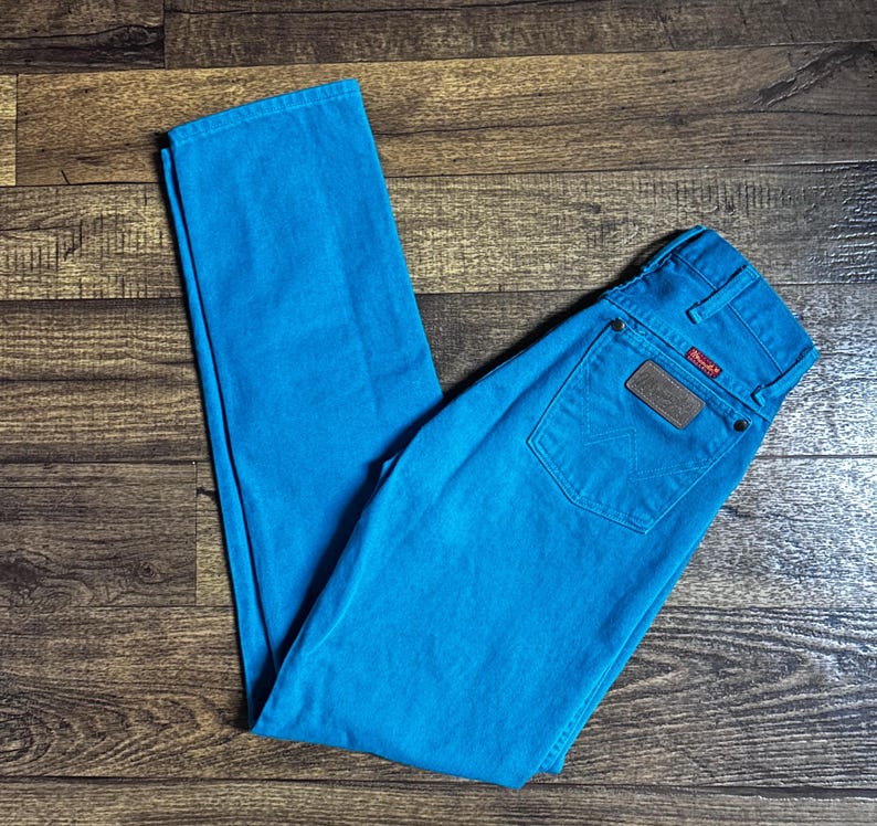 K&ouml;nnte beinhalten: Helle t&uuml;rkisfarbene Wrangler-Jeans. Die Jeans haben ein klassisches F&uuml;nf-Taschen-Design mit einem braunen Lederaufn&auml;her auf der Ges&auml;&szlig;tasche. Das Wrangler-Logo ist auf dem Aufn&auml;her sichtbar. Die Jeans liegen flach auf einer Holzoberfl&auml;che.