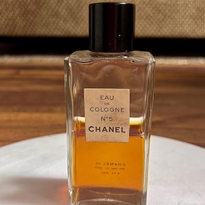 May include: A rectangular glass bottle of Chanel No. 5 Eau de Cologne. The bottle has a dark brown cap and a label with the text "EAU DE COLOGNE N&deg;5 CHANEL". The liquid inside is amber-colored.
