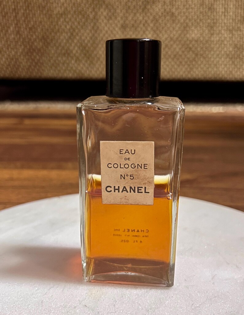 May include: A rectangular glass bottle of Chanel No. 5 Eau de Cologne. The bottle has a dark brown cap and a label with the text "EAU DE COLOGNE N&deg;5 CHANEL". The liquid inside is amber-colored.