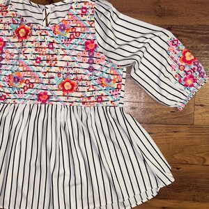 May include: White and black striped blouse with colourful floral embroidery on the front and sleeves.