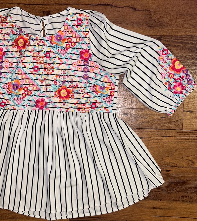 May include: White and black striped blouse with colourful floral embroidery on the front and sleeves.