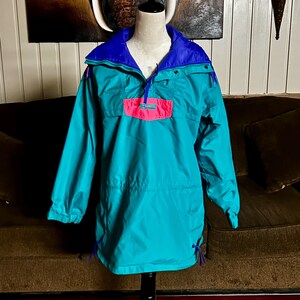 May include: A teal and blue anorak-style jacket with a pink pocket. The jacket has a drawstring waist and hood, and a half-zip closure. The jacket is displayed on a mannequin.