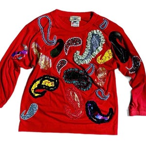 May include: A red long-sleeve shirt adorned with various paisley-shaped appliqués in diverse colors and textures. The appliqués include metallic gold, black, blue, and yellow designs, creating a unique and artistic look. The shirt has a crew neck and a tag.