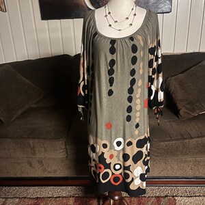 May include: A green, brown, black, white, and orange patterned dress with a scoop neck and 3/4 sleeves. The dress is on a mannequin and has a beaded necklace.