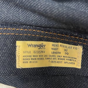 Może przedstawiać: Zbliżenie ciemnoniebieskich dżins&oacute;w z ż&oacute;łtą metką. Na metce widnieje napis "Wrangler" i zawiera numer stylu, rozmiar talii 81 cm i długość 76 cm. Dżinsy są wyprodukowane w USA.