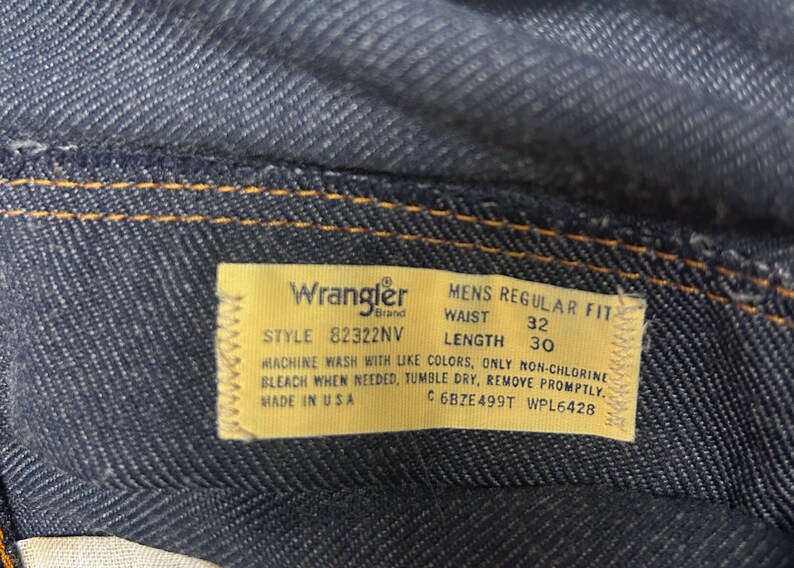 Może przedstawiać: Zbliżenie ciemnoniebieskich dżins&oacute;w z ż&oacute;łtą metką. Na metce widnieje napis "Wrangler" i zawiera numer stylu, rozmiar talii 81 cm i długość 76 cm. Dżinsy są wyprodukowane w USA.