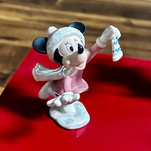 Może przedstawiać: Figurka Myszki Minnie w zimowym stroju. Figurka ma na sobie r&oacute;żowy płaszcz, białą czapkę i szalik. Trzyma małą, białą skarpetę z niebieskimi akcentami. Figurka stoi na białej podstawie.