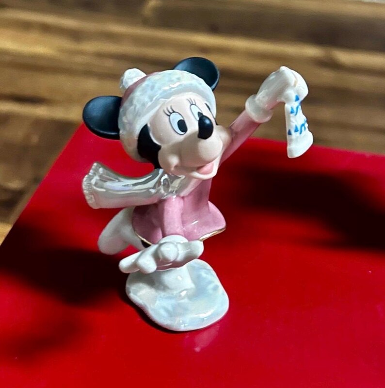 Może przedstawiać: Figurka Myszki Minnie w zimowym stroju. Figurka ma na sobie r&oacute;żowy płaszcz, białą czapkę i szalik. Trzyma małą, białą skarpetę z niebieskimi akcentami. Figurka stoi na białej podstawie.