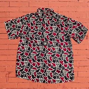 May include: A short-sleeved button-down shirt featuring a camouflage pattern in shades of maroon, black, and grey. The fabric is covered in a repeating white and black Texas Tech University logo. A small circular patch is centered on the back yoke.