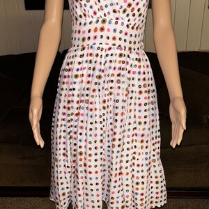 May include: White sleeveless dress with a V-neck and a fitted waist. The dress is adorned with a pattern of colorful polka dots in shades of blue, pink, orange, and brown. The dress falls to the knee.