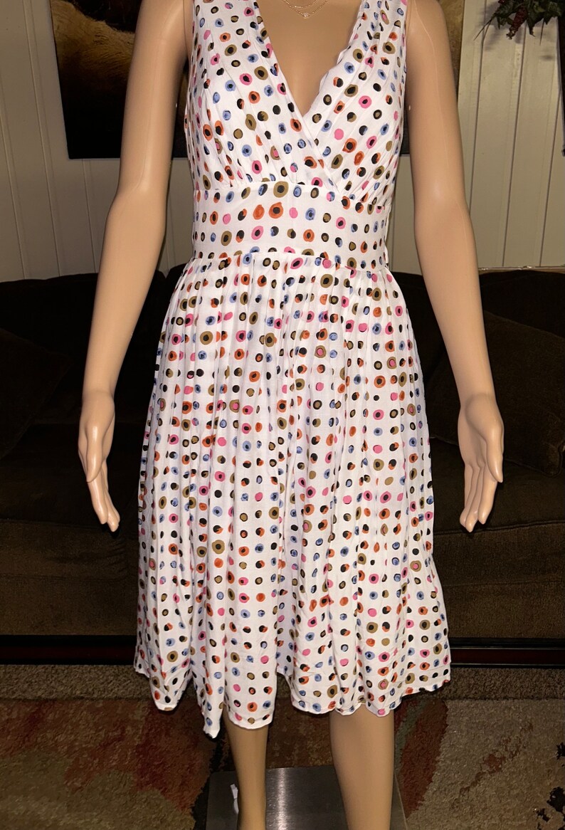 May include: White sleeveless dress with a V-neck and a fitted waist. The dress is adorned with a pattern of colorful polka dots in shades of blue, pink, orange, and brown. The dress falls to the knee.