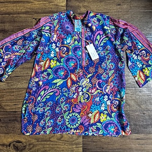May include: A vibrant, long-sleeved tunic with a colorful floral pattern in shades of blue, purple, pink, and green. The tunic features a half-zip neckline and wide sleeves with striped accents. A tag is attached to the tunic.