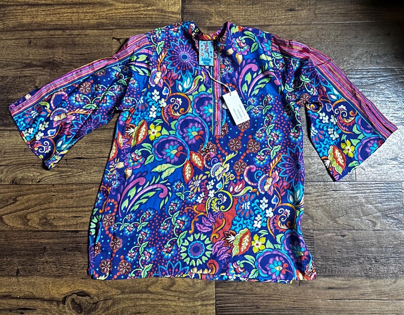 May include: A vibrant, long-sleeved tunic with a colorful floral pattern in shades of blue, purple, pink, and green. The tunic features a half-zip neckline and wide sleeves with striped accents. A tag is attached to the tunic.