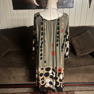 May include: A green, black, white, and orange patterned dress with a scoop neck and 3/4 sleeves. The dress is on a mannequin and is photographed against a brown couch.