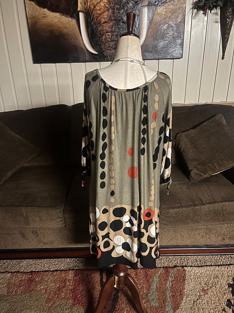 May include: A green, black, white, and orange patterned dress with a scoop neck and 3/4 sleeves. The dress is on a mannequin and is photographed against a brown couch.