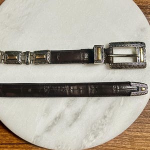 May include: A brown leather belt with silver and gold-toned metal accents. The belt features a rectangular buckle and decorative rectangular links. The leather has a textured appearance, and the metal has intricate patterns.