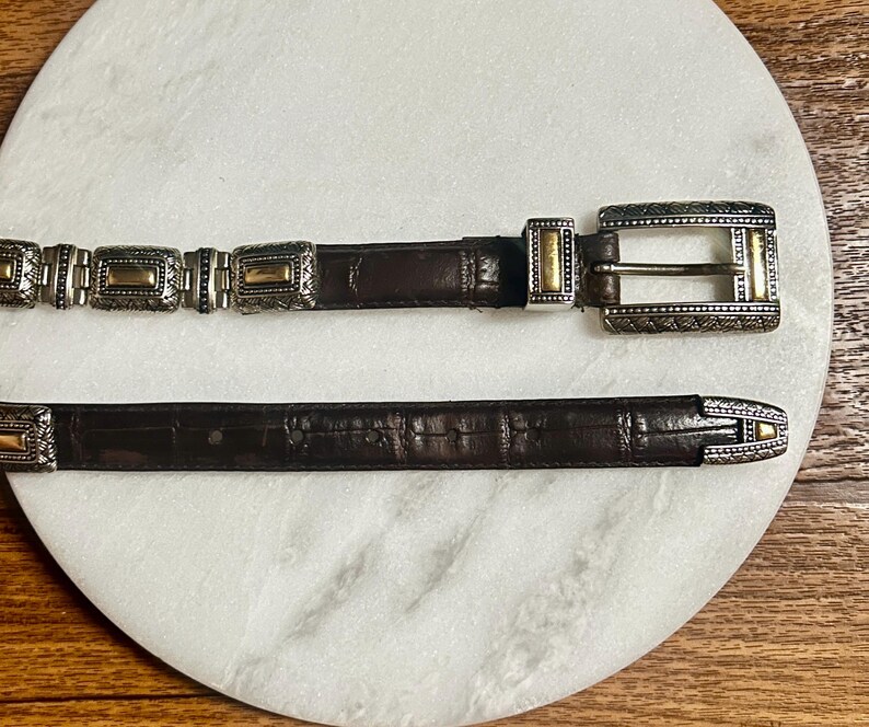 May include: A brown leather belt with silver and gold-toned metal accents. The belt features a rectangular buckle and decorative rectangular links. The leather has a textured appearance, and the metal has intricate patterns.