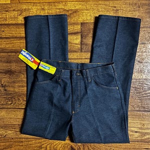 Może przedstawiać: Ciemnoniebieskie dżinsy Wrangler z klasycznym krojem. Dżinsy mają zapięcie na guziki, kieszenie i metkę Wrangler. Szwy są w kontrastowym ż&oacute;łtym kolorze. Dżinsy leżą płasko na drewnianej powierzchni.
