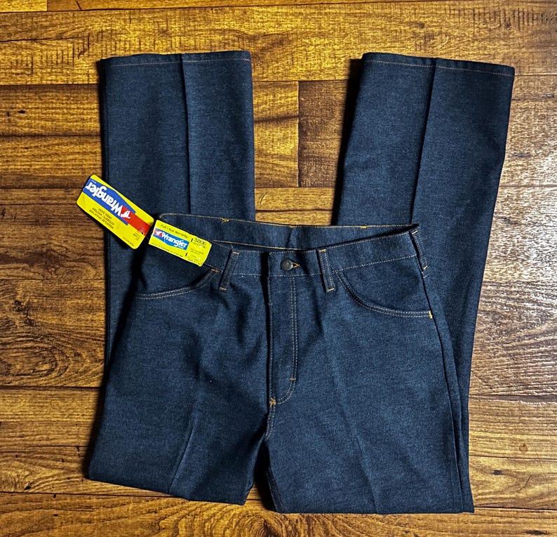 Może przedstawiać: Ciemnoniebieskie dżinsy Wrangler z klasycznym krojem. Dżinsy mają zapięcie na guziki, kieszenie i metkę Wrangler. Szwy są w kontrastowym ż&oacute;łtym kolorze. Dżinsy leżą płasko na drewnianej powierzchni.