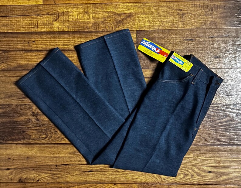 Może przedstawiać: Ciemnoniebieskie dżinsy Wrangler z denimu o prostym kroju nogawek. Dżinsy mają klasyczny styl z pięcioma kieszeniami i zapięciem na zamek błyskawiczny. Etykieta Wrangler jest dołączona. Dżinsy leżą na drewnianej powierzchni.