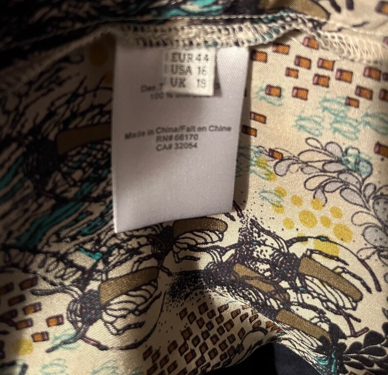 Puede incluir: Etiqueta blanca con texto negro que indica la talla EUR 44, USA 16, UK 15. La etiqueta tambi&eacute;n dice "Made in China/Fabriqu&eacute; en Chine" y "RN# 66170, CA# 32054". La etiqueta est&aacute; sobre una tela blanca con un patr&oacute;n floral negro y azul.