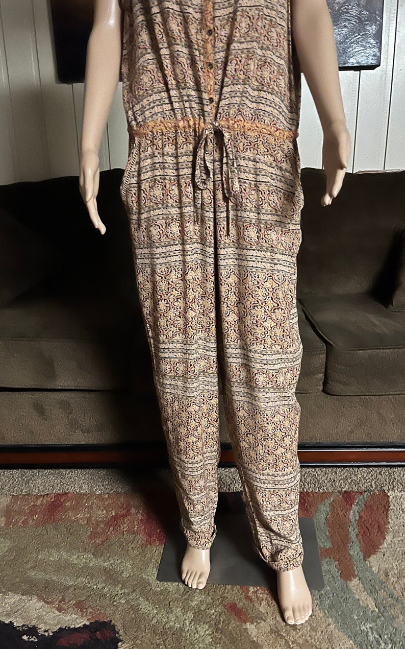 May include: A sleeveless jumpsuit with a beige background and a multi-colored floral pattern. The jumpsuit has a drawstring waist and a button closure at the front.