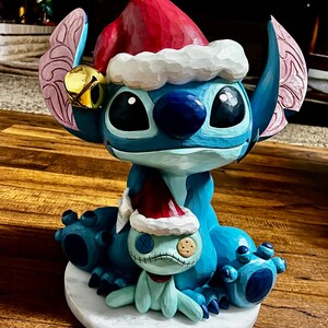 May include: A blue cartoon character figurine wearing a red Santa hat with white trim and a gold bell. The figurine is holding a smaller version of itself, also wearing a Santa hat. The figurine is sitting on a white marble base.